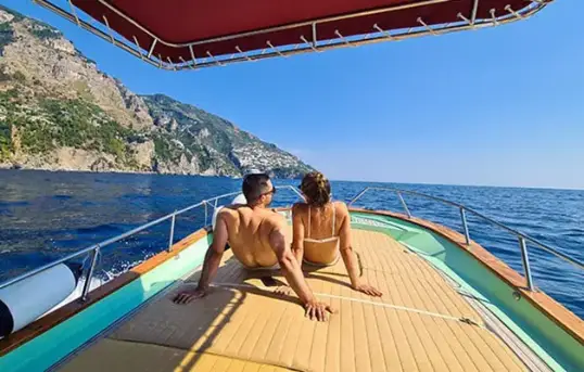 A relaxed couple sitting on the spacious bow sundeck of the traditional Gozzo Sorrentino boat Bellavita, cruising through the crystal clear waters of Praiano along the Amalfi Coast during a private half day boat excursion with Frasil Charter.